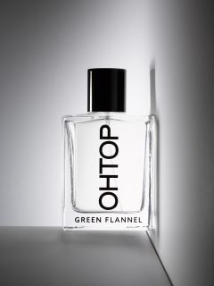 Ein durchsichtiger Parfümflakon mit der Aufschrift "OHTOP", einem schwarzen Verschluss und der Aufschrift "GREEN FLANNEL" am Boden, der an einer weißen Wand lehnt, über der "OH TOP" steht.