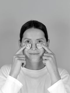 Eine Person in einem weißen Hemd lächelt, während sie zwei kleine, gebogene Objekte unter ihren Augen hält. Über ihrem Kopf erscheint das Wort "TEMPRASTONES".