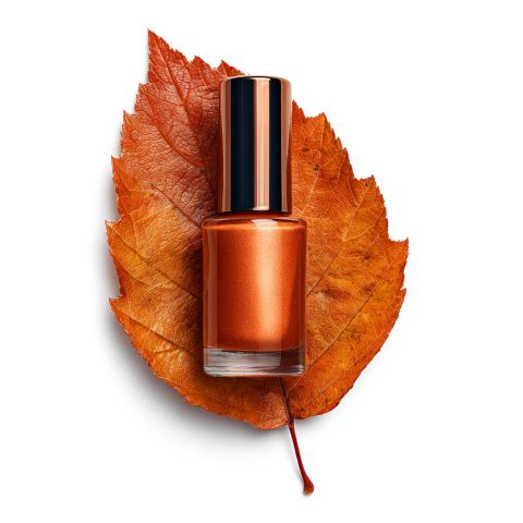Ein Fläschchen mit orangefarbenem Metallic-Nagellack steht auf einem getrockneten orangefarbenen Herbstblatt, beides vor einem weißen Hintergrund.