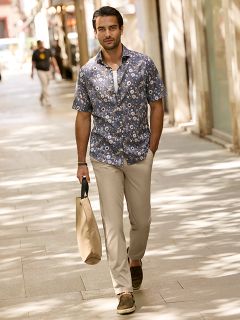 Ein Mann mit kurzen dunklen Haaren und einem Bart lehnt im Freien an einem steinernen Geländer. Er trägt eine rot-weiß karierte Jacke über einem cremefarbenen Pullover und blaue Jeans. Im Hintergrund sind städtische Gebäude zu sehen.