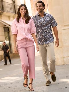 Eine Frau in einem gemusterten Pelzmantel und einem roten Oberteil steht lächelnd neben einem Mann in einer marineblauen Winterjacke und einem beigen Pullover. Sie stehen draußen an einer Steinmauer, und die Frau hält eine braune Handtasche in der Hand.