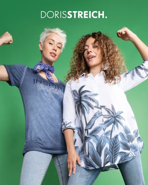 Zwei Frauen posieren selbstbewusst in schwarz-weißen Outfits mit grünen Akzenten, darunter eine Pufferweste, eine Mütze und Schuhe, vor einem dunklen Hintergrund mit dem Text "DORIS STREICH" über ihnen.