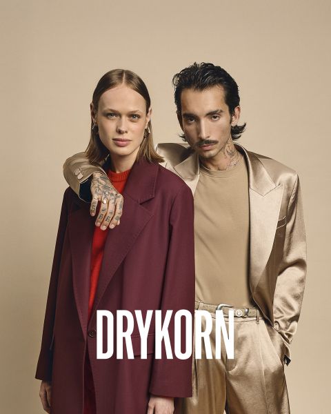 Eine elegante ältere Frau mit platinblondem Haar und einem schwarzen Pelzmantel sitzt auf einem alten Sofa neben einem großen braunen Hund. Das Wort "DRYKORN" ist am unteren Rand des Bildes zu lesen. Die warme Beleuchtung sorgt für eine luxuriöse Atmosphäre.