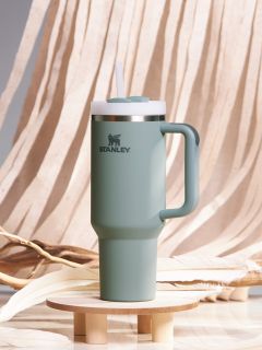 Ein grüner Stanley-Reisebecher mit Henkel und Strohdeckel steht auf einem kleinen Holzsockel. Der Hintergrund besteht aus fließendem beigem Stoff und dekorativen Zweigen.