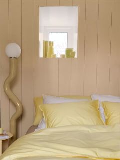 Ein ordentlich gemachtes Bett mit gelbem Bettzeug steht neben einer geschwungenen Stehlampe. Über dem Bett befindet sich ein kleines Fenster mit dekorativen Gegenständen und an der Wand steht "SUITE 702".