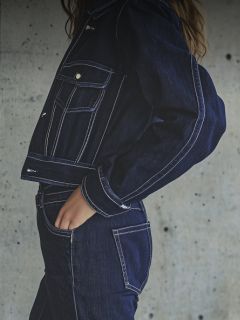 Eine Frau mit weißer Mütze, weißem T-Shirt und blauen Jeans posiert mit ausgestrecktem Arm in einem modernen, weißen Raum mit Blumen darüber.