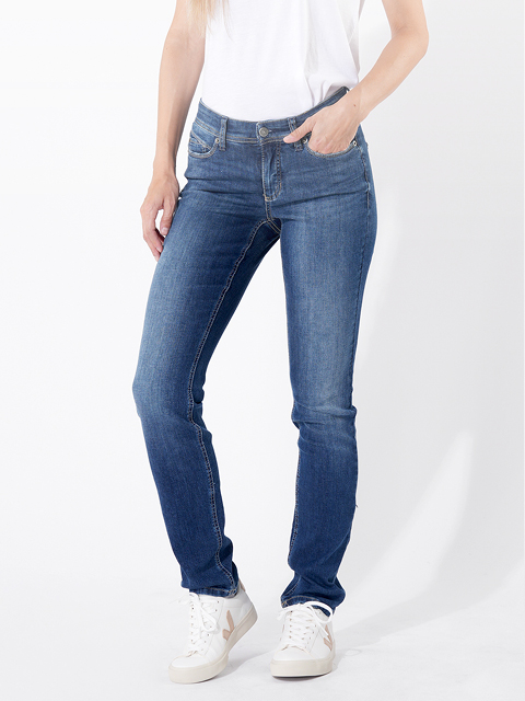Eine Person, die ein weißes T-Shirt, blaue Slim-Fit-Jeans und weiße Turnschuhe trägt, steht vor einem schlichten weißen Hintergrund und hat eine Hand in der Tasche.