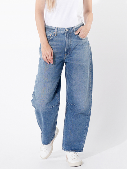 Eine Person, die eine locker sitzende blaue Jeans, ein weißes T-Shirt und weiße Turnschuhe trägt, steht mit einer Hand in der Tasche vor einem schlichten weißen Hintergrund.