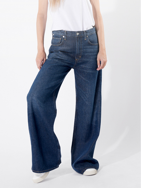 Eine Person, die eine weite Bluejeans und ein weißes T-Shirt trägt, steht vor einem einfarbig weißen Hintergrund. Nur die untere Hälfte des Körpers ist zu sehen; die Jeans ist weit geschnitten und verdeckt den größten Teil der Schuhe.