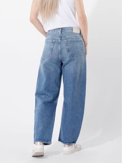 Eine Person, die eine locker sitzende blaue Jeans, ein weißes T-Shirt und weiße Turnschuhe trägt, steht mit einer Hand in der Tasche vor einem schlichten weißen Hintergrund.