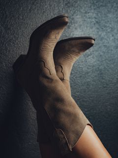 Nahaufnahme der Beine einer Person in taupefarbenen Cowboystiefeln aus Wildleder mit Ziernähten, die an einer strukturierten dunkelgrauen Wand lehnen.