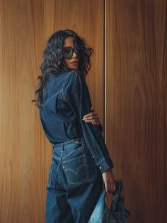 Eine Frau mit langem gewelltem Haar, Sonnenbrille und dunklem Jeans-Outfit steht vor einer Holzwand, schaut über die Schulter und hält eine Jeans-Tasche in der Hand.
