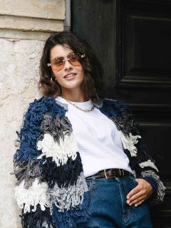 Eine Frau mit kurzen braunen Haaren, die eine Sonnenbrille, eine dicke blau-weiße Strickjacke, ein weißes T-Shirt und eine blaue Jeans trägt, steht lässig vor einer Steinmauer und einer dunklen Tür und lächelt selbstbewusst.