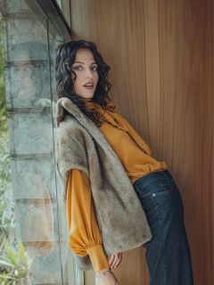 Eine Frau mit gewelltem dunklem Haar lehnt an einer Holzwand an einem Fenster. Sie trägt eine gelbe Bluse mit Puffärmeln, eine braune Fellweste und dunkle Jeans. Ihr Gesichtsausdruck ist entspannt, während sie in die Kamera blickt.