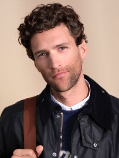 Ein Mann mit lockigem braunem Haar und blauen Augen posiert vor einem neutralen Hintergrund. Er trägt eine dunkle Jacke über einem Hemd mit Kragen und einem Pullover und hat ein braunes Band über der Schulter.