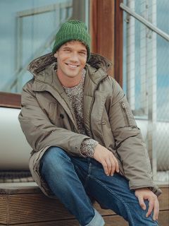 Ein lächelnder Mann, der eine grüne Strickmütze, einen grauen Wintermantel, blaue Jeans und einen gemusterten Pullover trägt, sitzt auf einer Holztreppe im Freien neben einem Metallgeländer.