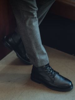 Person in grauer Hose und schwarzen Lederschnürstiefeln, ein Bein über dem anderen gekreuzt, steht in einem Innenraum auf einem hellen Boden.