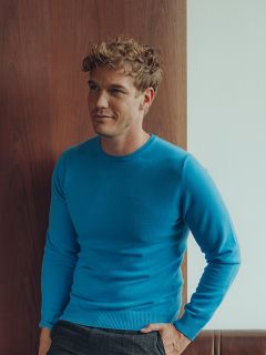 Ein Mann mit kurzen, lockigen blonden Haaren, der einen hellblauen Pullover und eine dunkle Hose trägt, steht drinnen an einer hölzernen und weißen Wand und blickt mit entspannter Miene leicht zur Seite.