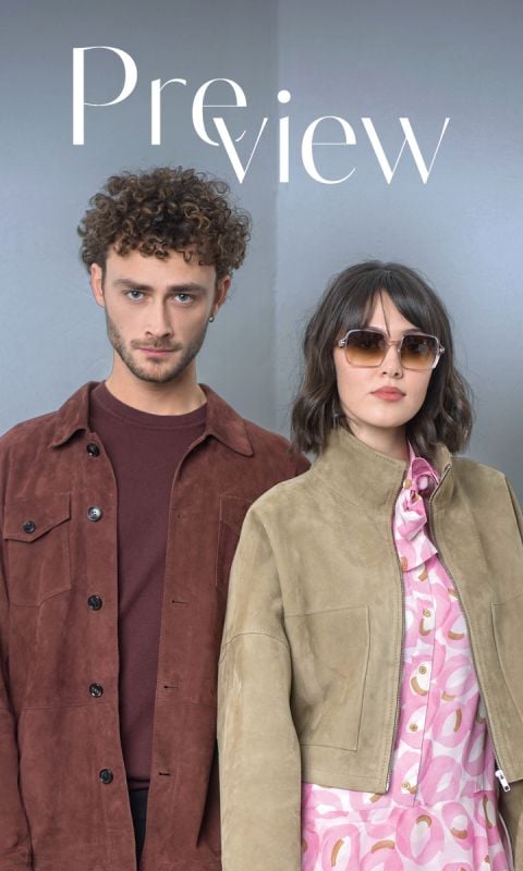 Ein Mann in einer braunen Jacke und eine Frau mit Sonnenbrille und hellbrauner Jacke über einem rosa gemusterten Kleid stehen nebeneinander vor einem schlichten hellen Hintergrund.