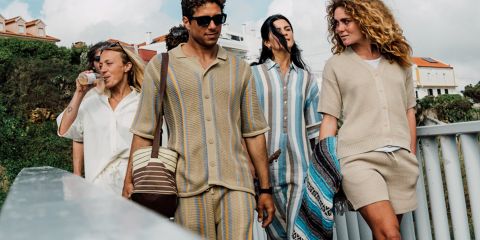 Eine Gruppe von fünf Personen macht draußen in der Nähe von Wasser ein Selfie, mit Bäumen und einem Sonnenuntergang im Hintergrund. Sie lächeln und posieren verspielt, in legerer Kleidung. Über ihnen sind der Text „KnowledgeCotton Apparel“ und ein Eulenlogo zu sehen.