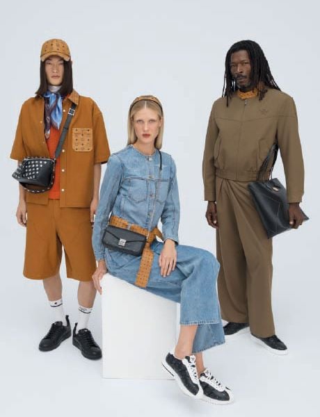 Eine blaue MCM-Designertasche mit weißem Logomuster steht auf hölzernen Stufen, die mit bunten Tier- und Objektschlüsselbändern geschmückt sind, darunter eine Rakete, ein Hase und ein Bär.