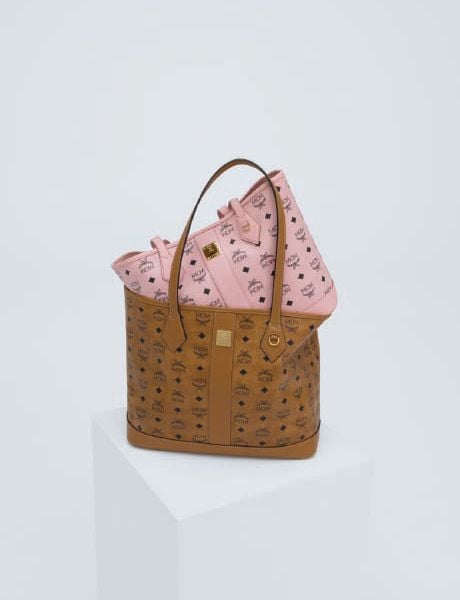 Eine Frau in einer hellbraunen Shearling-Jacke, schwarzen Shorts und Stiefeln steht auf einer Steinstufe und hält eine große Tasche. Im Hintergrund scheint ein Mann in bunter Kleidung waagerecht über den Stufen zu schweben. Hinter ihnen sind Bäume zu sehen.