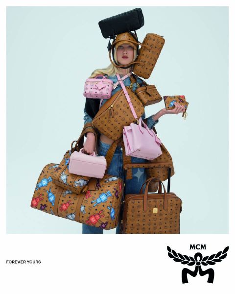 Drei Frauen in hellbraunen Mänteln und orangefarbenen Hüten gehen über eine Straße und tragen blaue MCM-Taschen mit einem weißen MCM-Logo im Vordergrund. Das Bild hat einen Spiegeleffekt, der die Szene dupliziert erscheinen lässt.