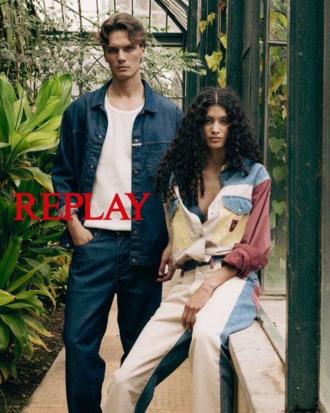 Ein Mann und eine Frau sitzen in dunkler, stilvoller Kleidung auf einer Holztreppe in einem Innenraum. Das Wort "REPLAY" ist in der Mitte des Bildes in roten Großbuchstaben prominent eingeblendet.