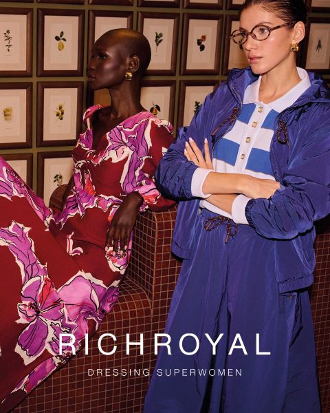 Eine Frau in hellblauem Hemd und grauer Hose hält sich eine Schneekugel vor ein Auge. Sie posiert in einem Raum mit Holzwänden und einem Skiplakat und trägt große Ohrringe. Der Text lautet "RICHROYAL DRESSING SUPERWOMEN".