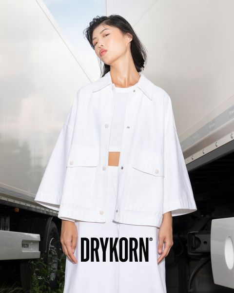 Eine Person steht selbstbewusst vor einem weißen LKW. Sie trägt eine lockere weiße Jacke und Hose, darunter ein DRYKORN-Logo. Im Hintergrund sind die Seite des LKWs und ein teilweise bewölkter Himmel zu sehen.