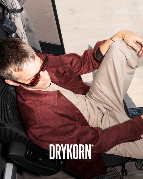 Eine Person mit dunkler Sonnenbrille und burgunderfarbener Jacke sitzt entspannt in einem Fahrzeug. Sie hat ihre Beine auf einem anderen Sitz hochgelegt und trägt beige Hosen und ein helles Hemd. Unten ist deutlich das Wort „DRYKORN“ zu sehen.