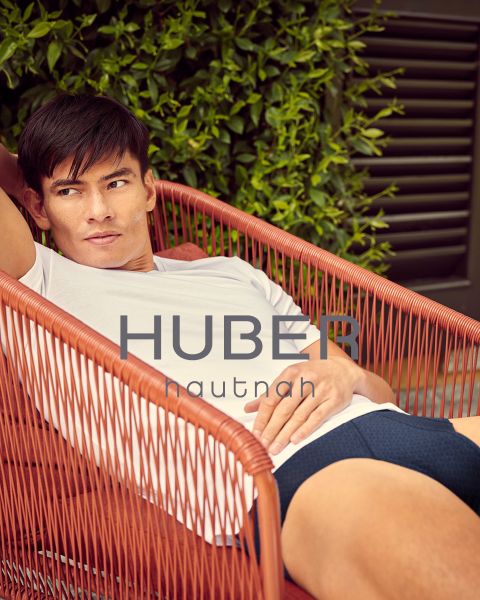 Ein lächelnder Mann in einem weißen gerippten Tanktop und Unterwäsche steht in einem Badezimmer mit warmer Beleuchtung, einem Spiegel und runden Lichtern hinter ihm. Der Text "HUBER hautnah" wird am unteren Rand des Bildes angezeigt.