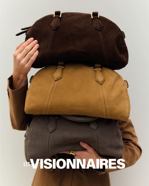 Eine Person in einem braunen Mantel hält eine Hand hoch, um das Gesicht teilweise zu verdecken. Sie steht vor einem dunklen Hintergrund. Oben im Bild sind in fetten weißen Buchstaben die Worte „Les Visionnaires“ geschrieben.