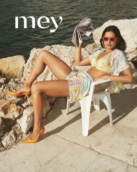 Ein Mann in schwarzen Boxershorts und eine Frau in einem weißen Tanktop und Unterwäsche stehen nebeneinander im Freien an einem Geländer, mit dem Meer und dem bewölkten Himmel im Hintergrund. Das Wort "mey" steht am unteren Rand.