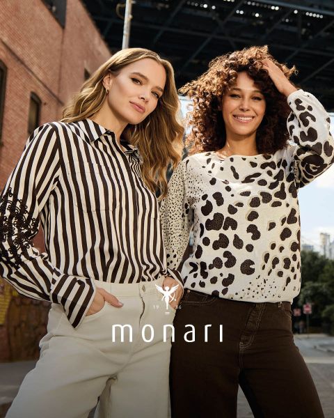 Eine lächelnde Frau, die einen beigen Mantel, ein gestreiftes Hemd, blaue Jeans und einen hellen Schal trägt, steht im Freien vor einem hohen Gebäude. Das Monari-Logo ist am unteren Rand des Bildes zu sehen.