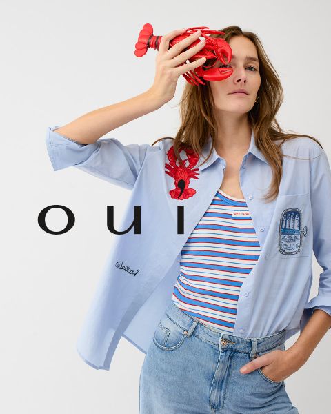 Eine Frau, die einen roten Pullover mit einem weißen Herz, blaue Jeans und eine weiße Mütze trägt, posiert vor einer hellen Wand. Am unteren Rand des Bildes steht in weißen Buchstaben das Wort "oui".