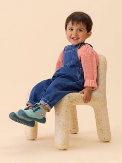 Ein Kleinkind in gestreiftem Hemd und Jeans-Overall sitzt mit einem Bein im Freien und zeigt die strukturierte blaue Sohle seines Schuhs.