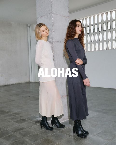 Eine Frau in einem weißen bauchfreien Top und beigen Hosen mit orangefarbenen Akzenten sitzt auf einem Hocker. Sie befindet sich in einem rustikalen Raum mit großen Fenstern. In der Mitte ist der Text „ALOHAS“ zu sehen.