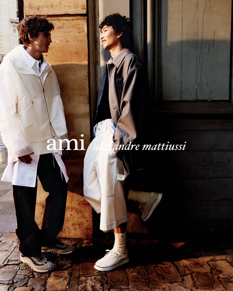 Drei eng beieinander stehende Personen in stilvollen Outfits, eine hält ein Getränk in der Hand. Der Text "ami alexandre mattiussi" ist in weißen Buchstaben über das Bild gelegt. Ihre Gesichter sind nicht zu sehen.