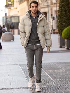 Ein Mann mit kurzen dunklen Haaren und einem Bart lehnt im Freien an einem steinernen Geländer. Er trägt eine rot-weiß karierte Jacke über einem cremefarbenen Pullover und blaue Jeans. Im Hintergrund sind städtische Gebäude zu sehen.