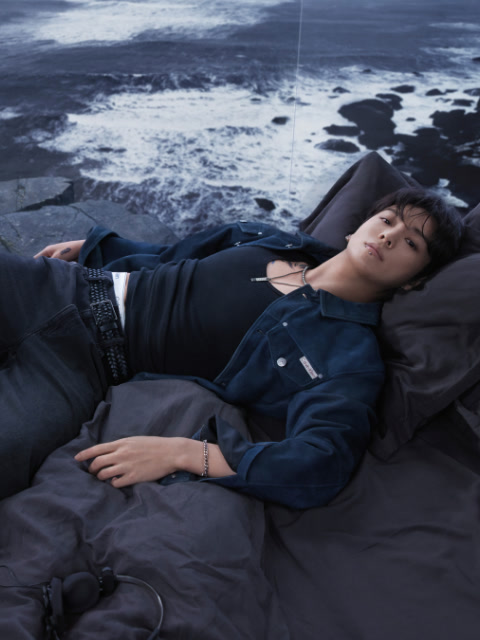 Schwarz-Weiß-Foto einer Frau in einem Calvin Klein Sport-BH und Jeans, die auf dem Rücken liegt, die Beine über den Kopf angewinkelt, die Arme auf dem Boden ausgestreckt, den Blick in die Kamera gerichtet.