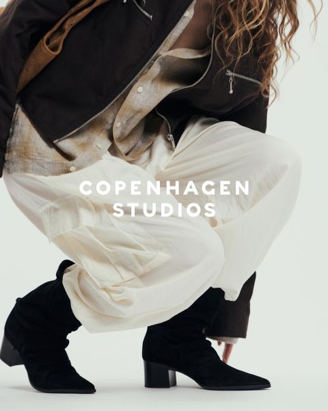 Eine Person in klobigen schwarzen Schnürstiefeln und dunklen Hosen steht auf einer weißen Fläche. Ein Teil des Bildes wird durch rosa und beigefarbenes, unscharfes Garn verdeckt. Der Text "Copenhagen Studios" erscheint über dem Foto.
