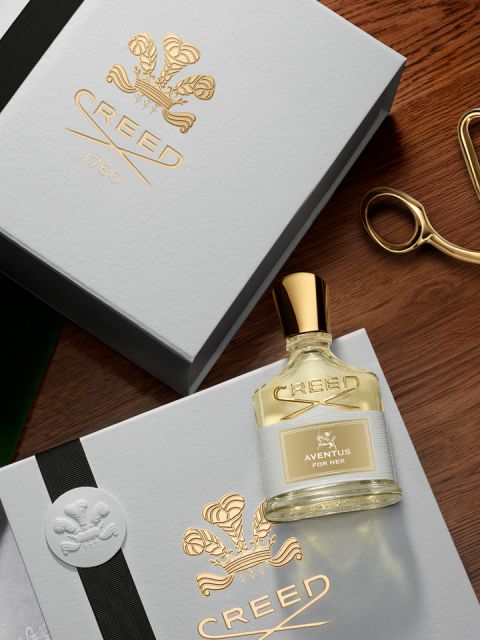 Eine rosa Flasche Creed Carmina-Parfüm steht vor einem rosa nebligen Hintergrund und ruht auf einer Oberfläche, die rosa Sand ähnelt. Die Flasche hat einen goldenen Verschluss und zeigt den Markennamen und das Logo in erhabener Schrift.