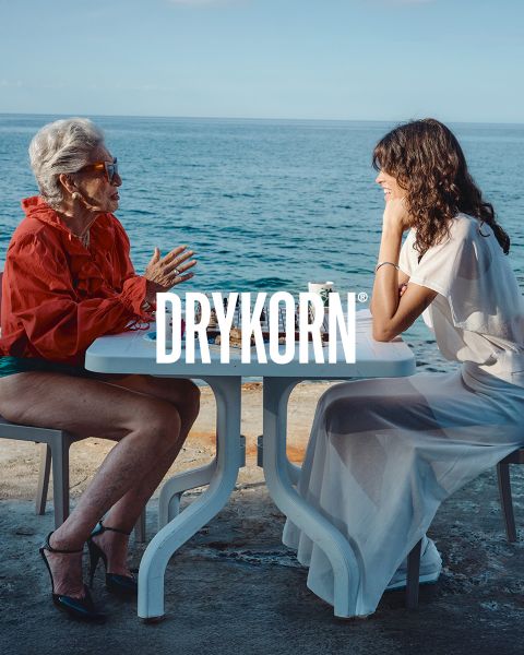 Eine elegante ältere Frau mit platinblondem Haar und einem schwarzen Pelzmantel sitzt auf einem alten Sofa neben einem großen braunen Hund. Das Wort "DRYKORN" ist am unteren Rand des Bildes zu lesen. Die warme Beleuchtung sorgt für eine luxuriöse Atmosphäre.