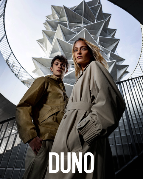 Eine Person steht im Profil, trägt eine hellgelbe Jacke und einen weißen Rock, hat den Kopf nach hinten geneigt und die Augen geschlossen. Sie befinden sich in einer Außenumgebung mit großen Kakteen und Palmen. Das Wort „DUNO“ erscheint vertikal auf der linken Seite des Bildes.