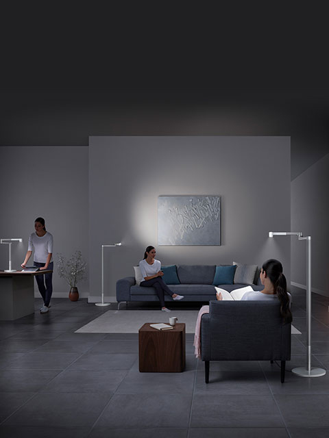 In einem schwach beleuchteten Wohnzimmer sind drei Personen mit unterschiedlichen Tätigkeiten beschäftigt: eine steht an einem Schreibtisch, eine andere sitzt mit einem Buch auf einem Sofa und eine dritte sitzt lesend auf einem Stuhl. Modernes Dekor und Stehlampen erhellen den Raum.