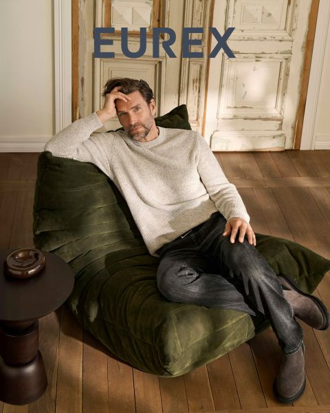 Ein Mann steht im Freien in der Nähe von hohem Gras, trägt einen beigen Pullover, eine marineblaue Pufferweste und eine braune Hose und schaut zur Seite. Das "EUREX by BRAX"-Logo erscheint auf der rechten Seite. Der Himmel ist bewölkt.