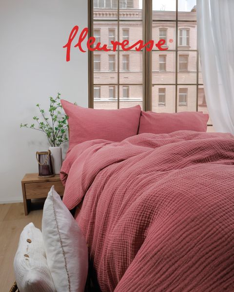 Ein gemütliches Schlafzimmer mit einem ordentlich gemachten Bett mit weißer Bettwäsche mit Vogelmuster. Zwei dekorative Kissen liegen am Kopfteil. Sonnenlicht fällt durch transparente Vorhänge und wirft einen sanften Schein auf den hellen Holzboden.