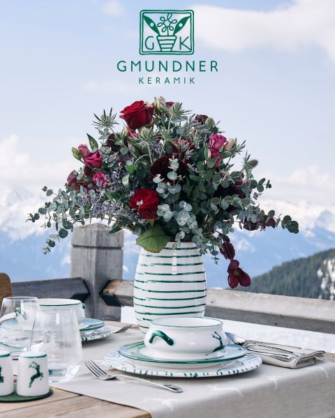 Ein wunderschön gedeckter Tisch mit dekorativen Keramiktellern mit abstrakten schwarzen Linienmustern. Eukalyptusblätter liegen über die Teller. Eine goldene Gabel, ein gestreiftes Sahnekännchen und ein gemusterter Becher ergänzen die elegante rosa Tischdecke.