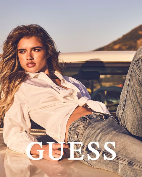 Zwei Frauen stehen dicht beieinander im Freien und tragen strukturierte Mäntel, eine in hellbraun und eine in cremefarben. Beide halten ihren Kragen fest und schauen nach vorne. Das Wort "GUESS" steht in fetten weißen Buchstaben am unteren Rand.